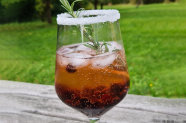 Mit rötlicher Flüssigkeit, Beeren und Eiswürfeln gefülltes Weinglas mit Zuckerrand, aus dem Rosmarin ragt