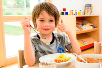 Kleiner Junge sitzt am Kita Esstisch mit einem Teller Essen vor sich und Löffel in der Hand.